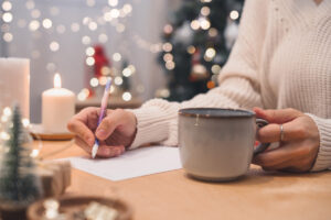 A hand writes with a pen and holds a mug, with Christmas lights in the background.