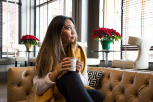A woman is sitting on the couch, holding a mug, and gazing out of a window in a cozy home setting.