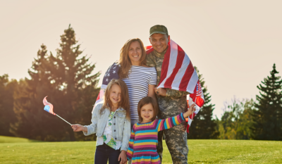 Military family smiling outdoors, with the service member in uniform holding an American flag.