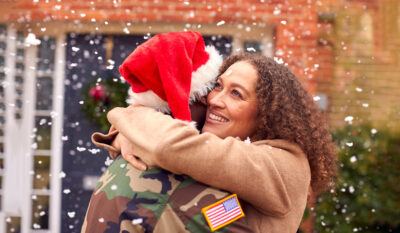 Military spouse hugging service member with holiday hat with snow falling
