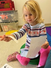 Two-year-old military child seated on a floor pad, sharing her papers from Operation Little Learners in wonderment.