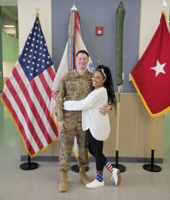 Military spouse Kalyn hugging her service member husband with flags behind them